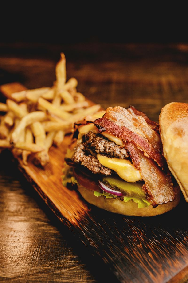 Juicy cheeseburger stacked with leftover BBQ meat, bacon, cheese, pickles, lettuce, and onions served with golden fries on a wooden board.