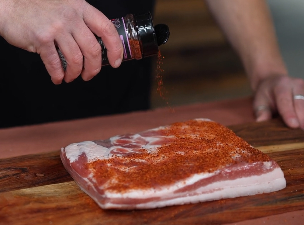 This image shows pork belly being seasoned with Boomas BBQ Swine & Dine rub