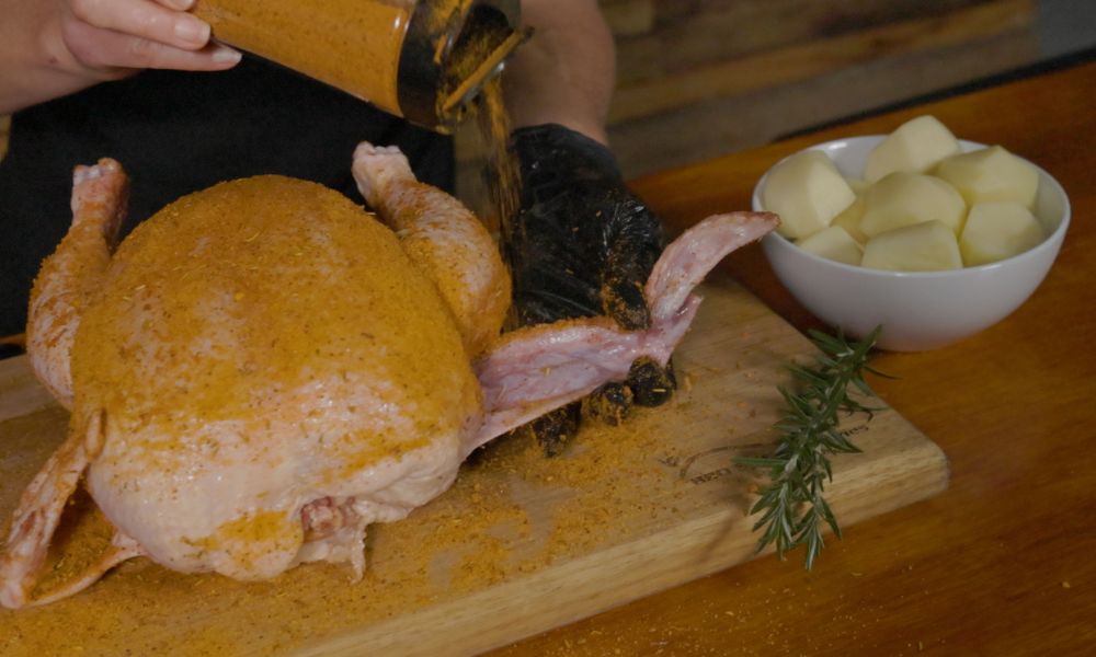 Whole chicken being seasoned with Flaming Coals Clucking Mad rub, next to peeled potatoes and rosemary on a wooden board.