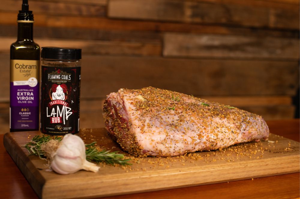 Raw leg of lamb seasoned with Flaming Coals Lamb Rub, displayed on a wooden board with garlic, rosemary, and olive oil.