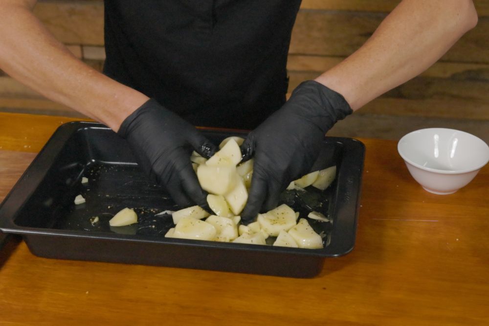Seasoning and mixing chopped potatoes in a black baking tray, preparing them for roasting.