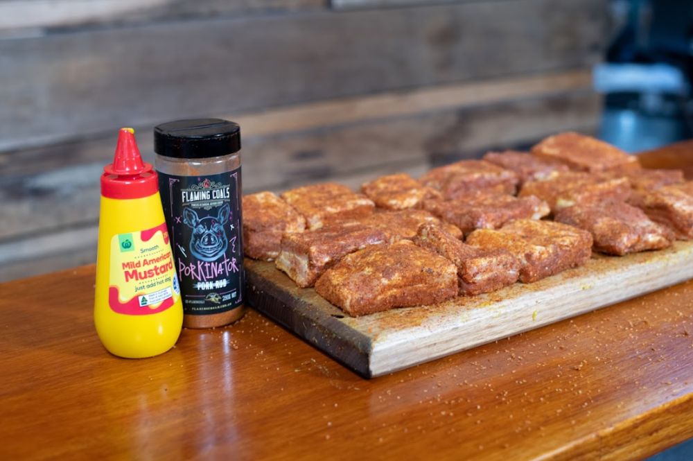 Seasoned pork belly cubes resting on a wooden board, coated in dry rub. A bottle of  American mustard and a jar of Flaming Coals Porkinator rub are placed beside the pork, showing the ingredients used for flavouring.