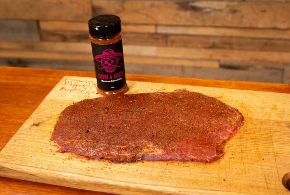 Raw skirt steak coated with a dry spice rub on a wooden cutting board, with a bottle of Booma’s BBQ "Fiesta & Siesta" Mexican seasoning placed behind it.