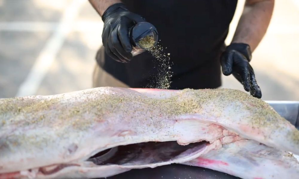 Whole lamb being coated with seasoning on a stainless steel prep tray, ready for spit roasting.