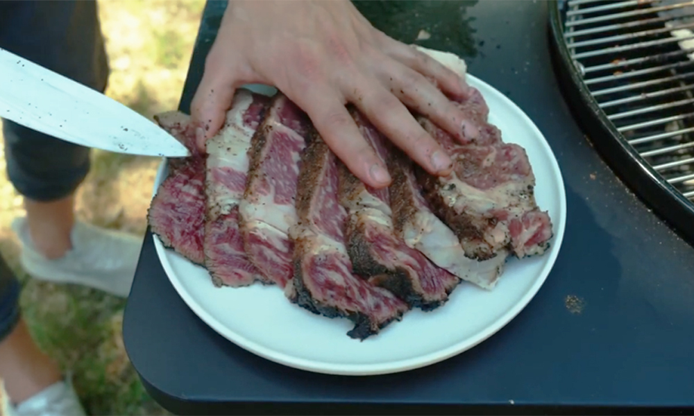 Sliced steak served on a white plate beside the SNS Master Kettle Grill.