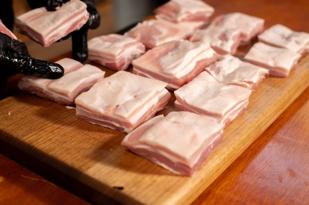 Raw pork belly cubes arranged on a wooden cutting board, with a person wearing black gloves placing one piece. The pork belly shows distinct layers of meat and fat, ready for seasoning or cooking.