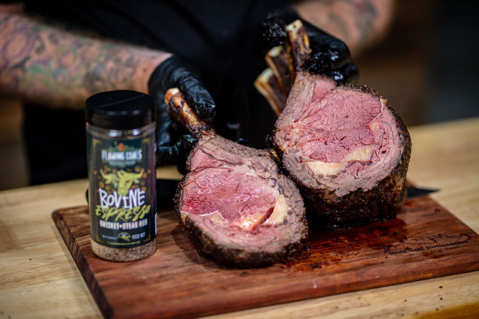Juicy ribeye roast cut in half, showing a tender medium-rare centre with a seared crust, placed on a wooden chopping board. A jar of Flaming Coals Bovine Espresso Brisket & Steak Rub is in the foreground, with a person wearing black gloves holding the meat.