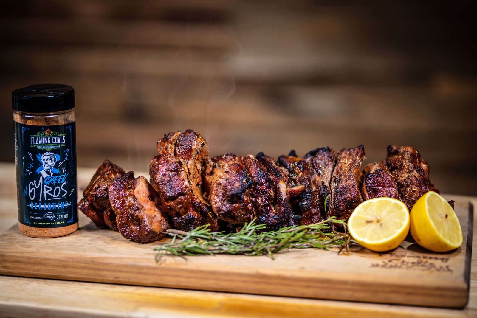 Juicy roast beef slices cooked on a spit, served on a wooden board with fresh rosemary and halved lemons. A jar of Flaming Coals Greek Gyros Seasoning is placed beside the meat.