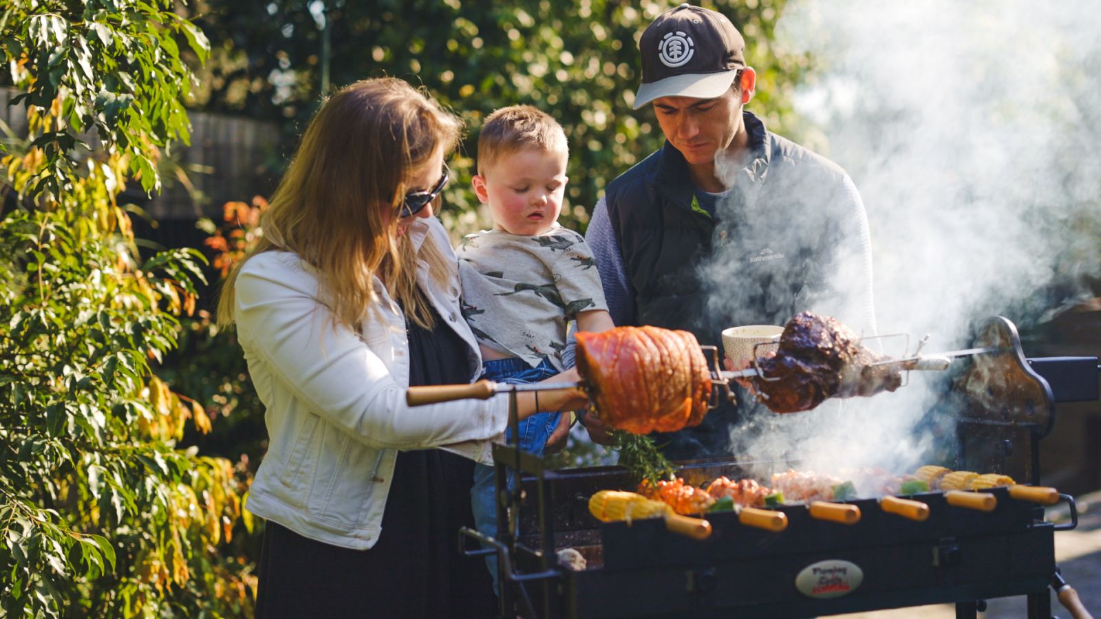 Family cooking together outdoors on a Flaming Coals Cyprus Spit, roasting meats and vegetables over charcoal.
