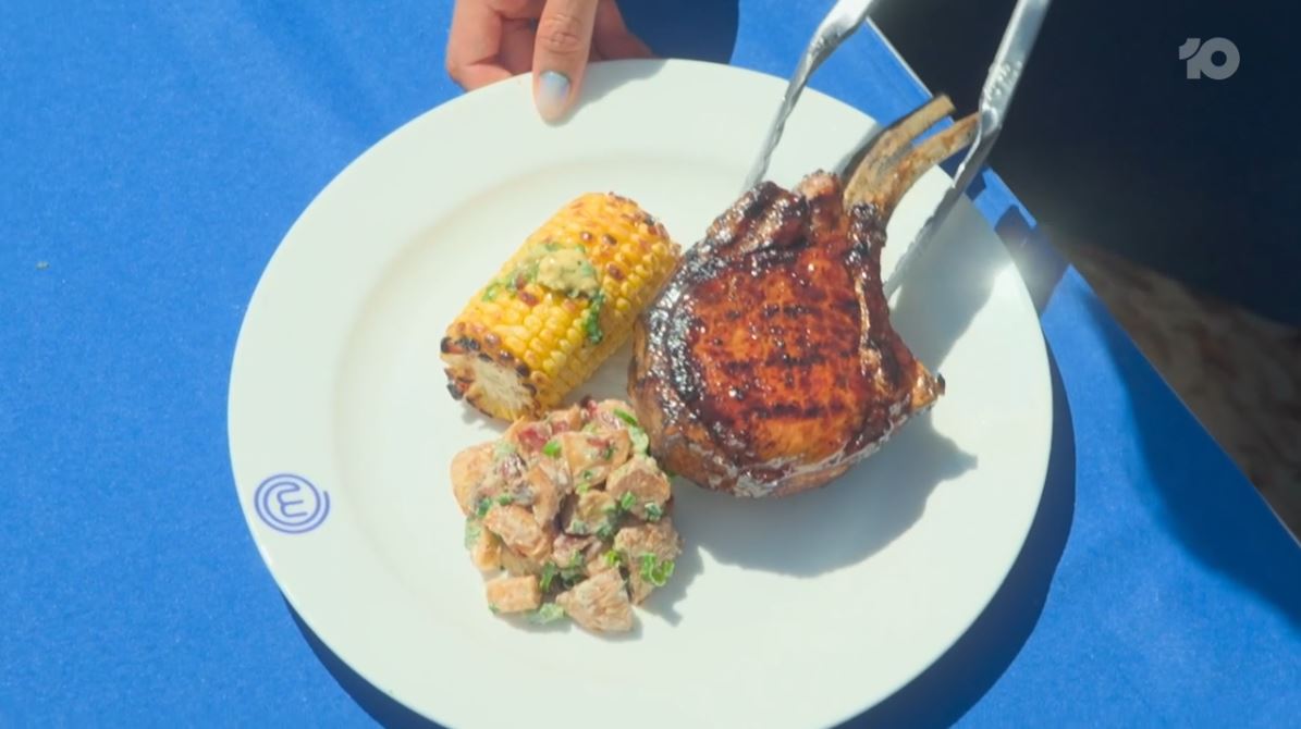 Plated meal featuring a grilled bone-in steak, roasted corn on the cob, and potato salad, with tongs holding the steak, set against a blue background.