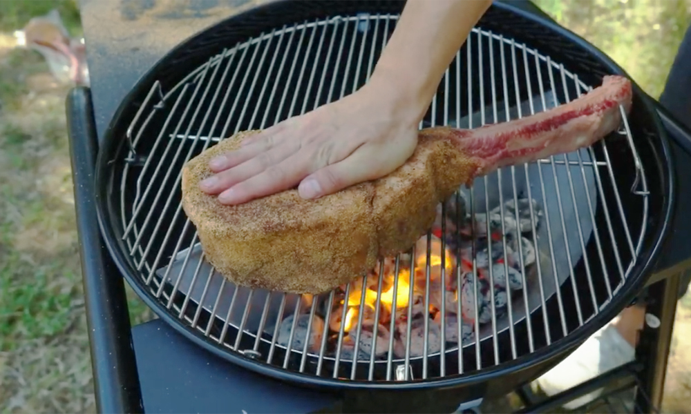Seasoned tomahawk steak being placed on the grill over hot coals in an SNS Master Kettle BBQ.