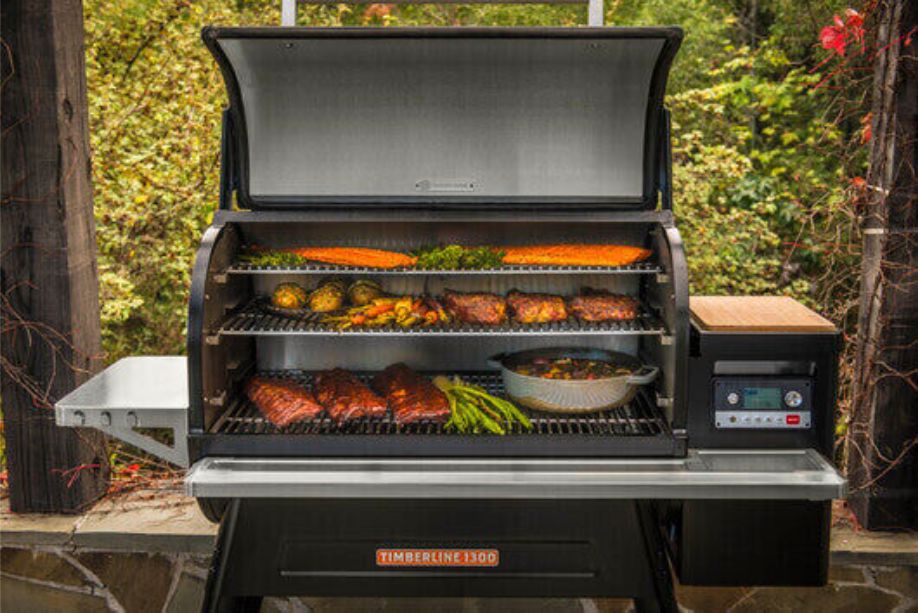 Traeger Timberline 1300 BBQ Meat Smoker with open lid showing multiple racks filled with ribs, salmon, vegetables, and a pan of food, set outdoors on a patio with trees in the background.