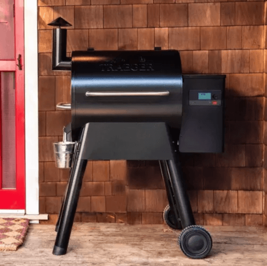 Traeger Pro 575 Pellet Grill in black, set up on a wooden deck outside a home with red door and shingle siding.