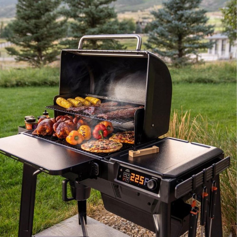 Traeger Woodridge pellet grill cooking chicken, corn, steak and vegetables with the lid open, showing digital temperature controller and side shelves in a backyard setting.