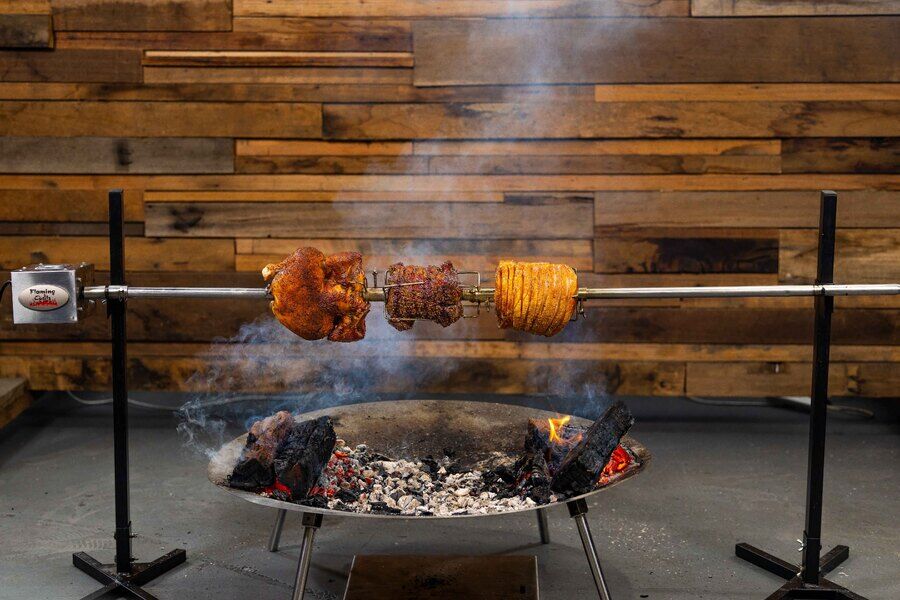 Portable camping spit roasting pork and beef over an open fire pit using a Flaming Coals rotisserie setup.