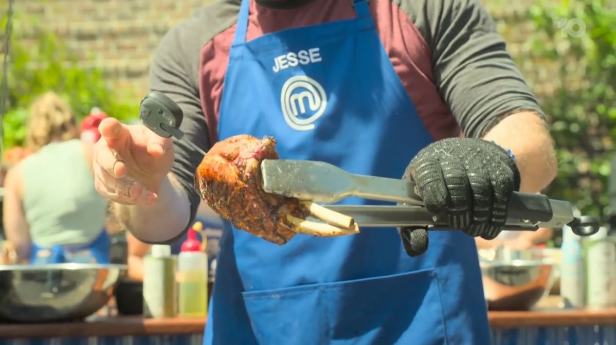 MasterChef contestant, wearing a blue apron, using tongs to check the temperature of a cooked steak while holding a probe thermometer, with other contestants working in the background.