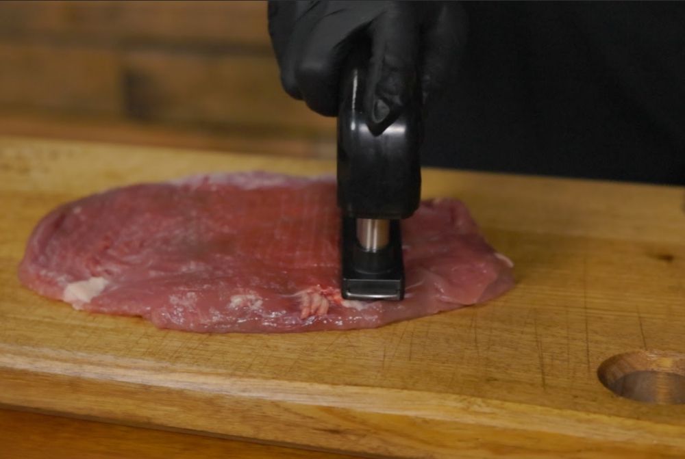 A person wearing black gloves uses a meat tenderiser tool to tenderise a raw skirt steak on a wooden cutting board.