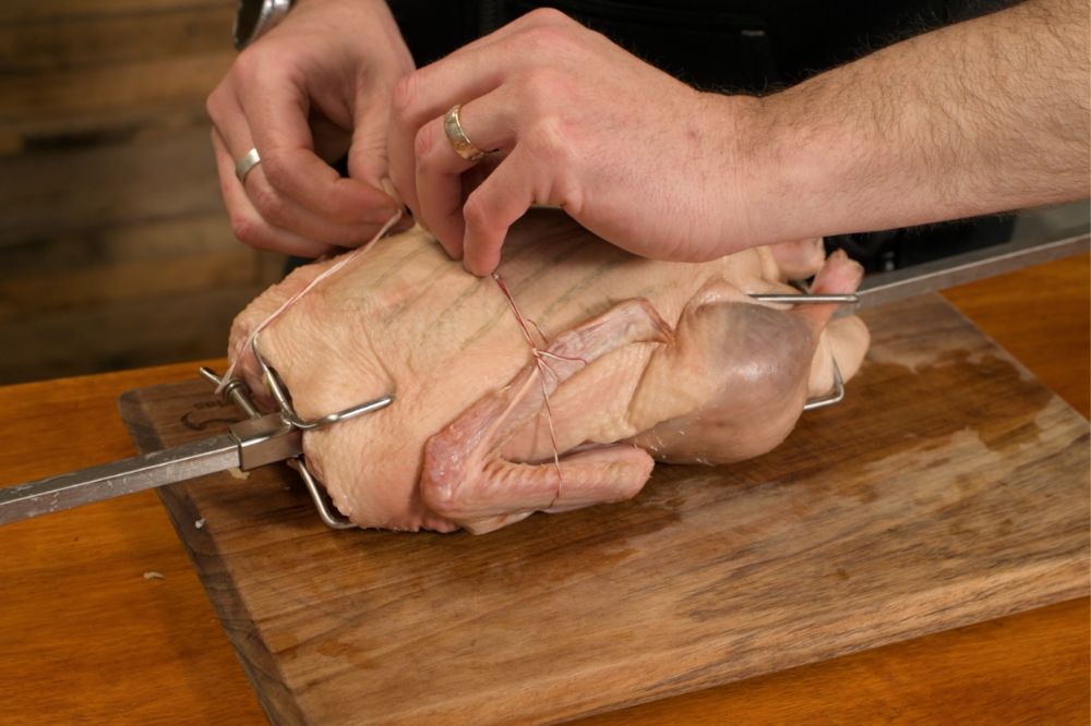 Whole duck secured on a spit rod with butcher’s twine, wings and legs tied in place, and stuffed with ingredients for spit roasting.