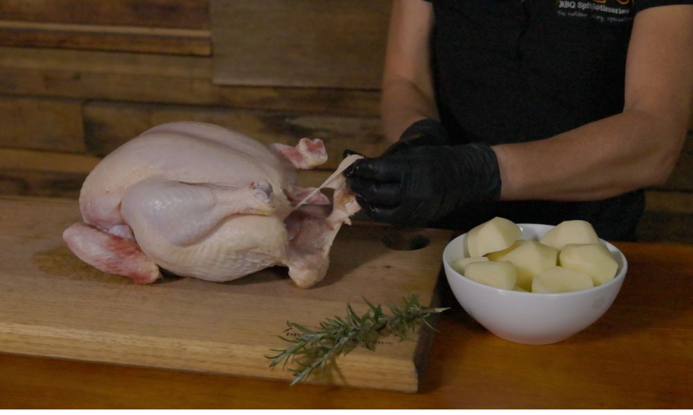  A person wearing black gloves trims excess fat from a raw whole chicken on a wooden cutting board, with a bowl of peeled potatoes and fresh rosemary nearby.
