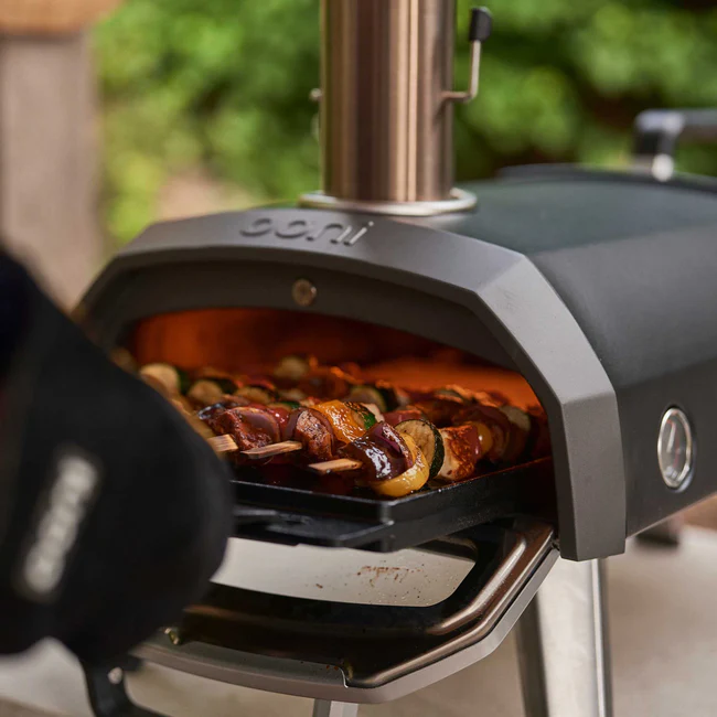 Close-up of the Ooni Karu 2 Multi-Fuel Pizza Oven in use, with skewers of grilled vegetables and meat roasting inside on a tray. A person wearing a heat-resistant glove is handling the food while the oven’s interior glows with heat, set outdoors against a blurred garden background.