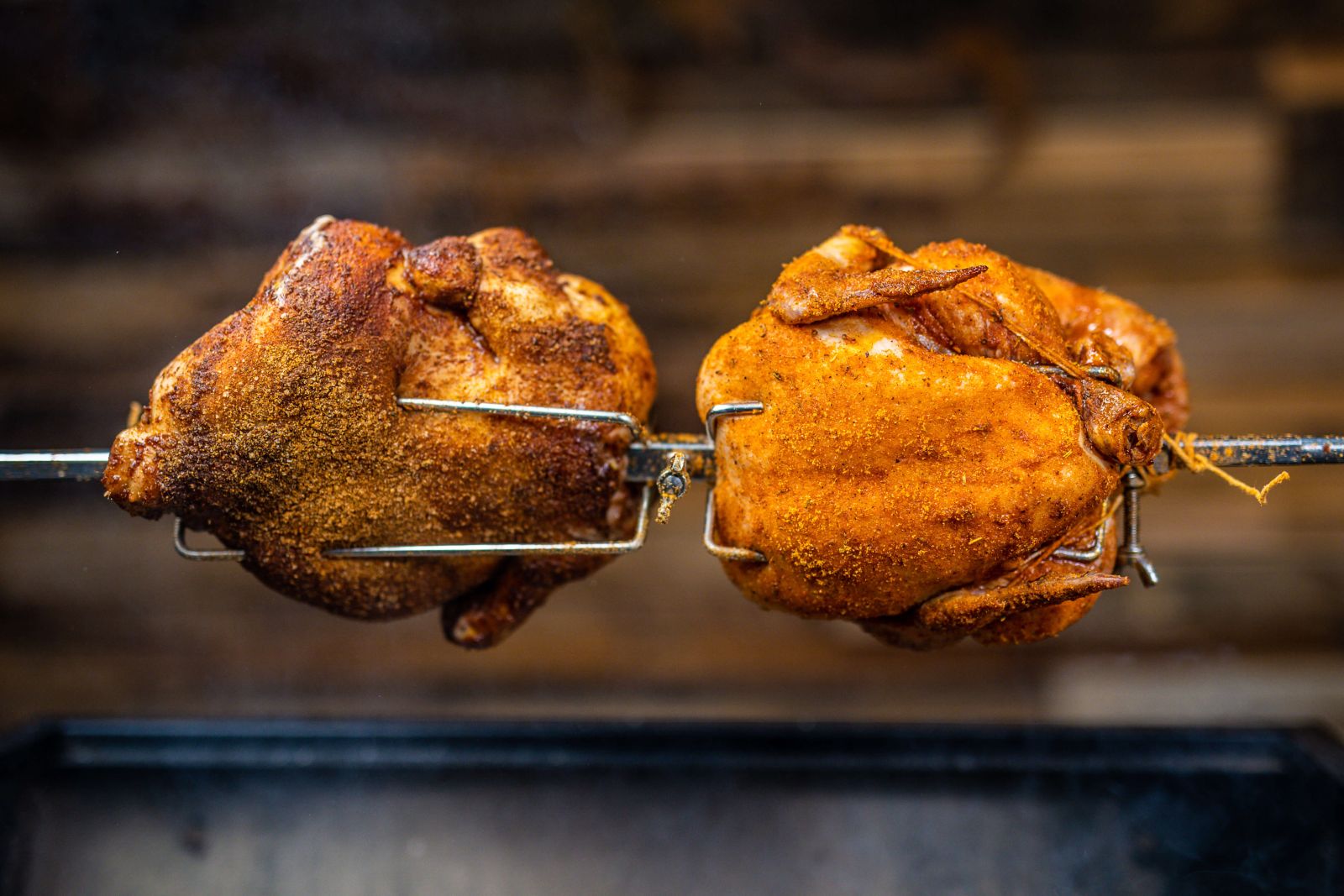 Close-up of two whole chickens roasting on a rotisserie spit with seasoning.