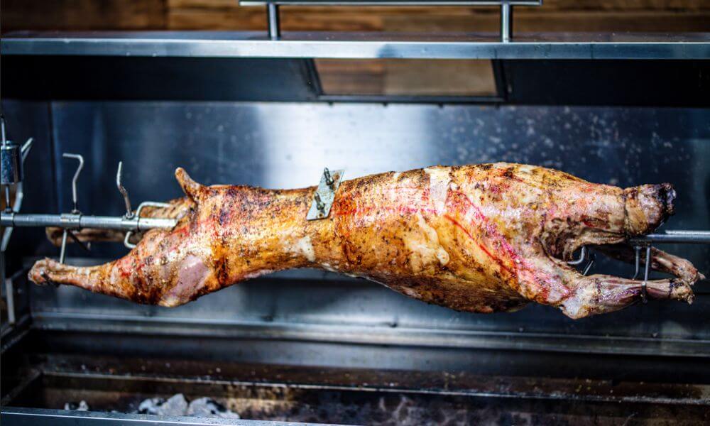 Whole lamb roasting on a stainless steel spit roaster with counter balance weight over charcoal.