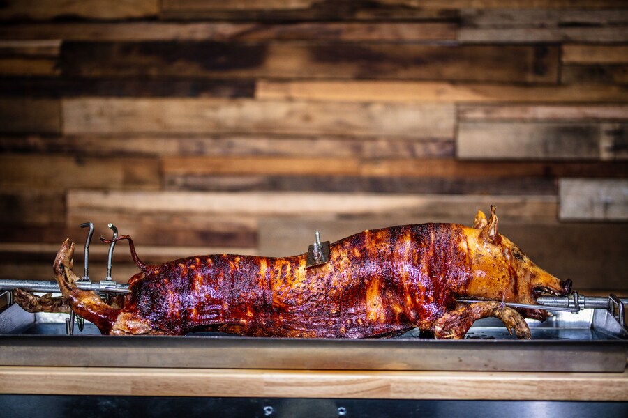 Whole pig roasted on a stainless steel rotisserie spit, golden brown with crispy skin and an inserted meat thermometer, set against a wooden wall background.