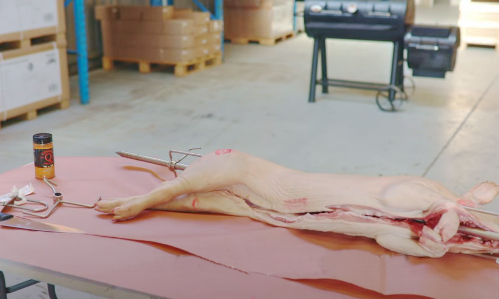 Whole raw pig laid out on a table with a rotisserie skewer inserted, ready for preparation. A jar of seasoning and spit roasting accessories are placed nearby, with a smoker grill visible in the background.