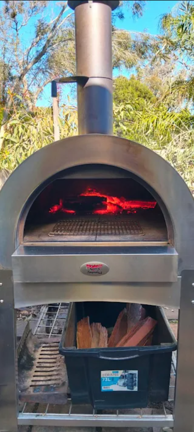 Outdoor wood fired pizza oven with burning red-hot embers inside, chimney above, and a crate of firewood stored below