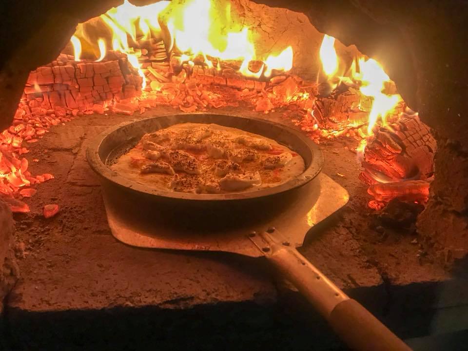 Aluminium-head pizza peel sliding a pizza into a wood-fired oven, surrounded by glowing coals and flames.
