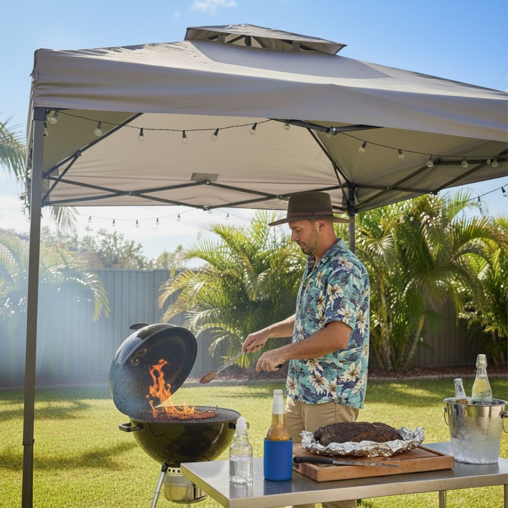 Man barbecuing under a backyard gazebo on a sunny day, tending a charcoal grill with flames while a cooked roast rests on a table beside chilled drinks.