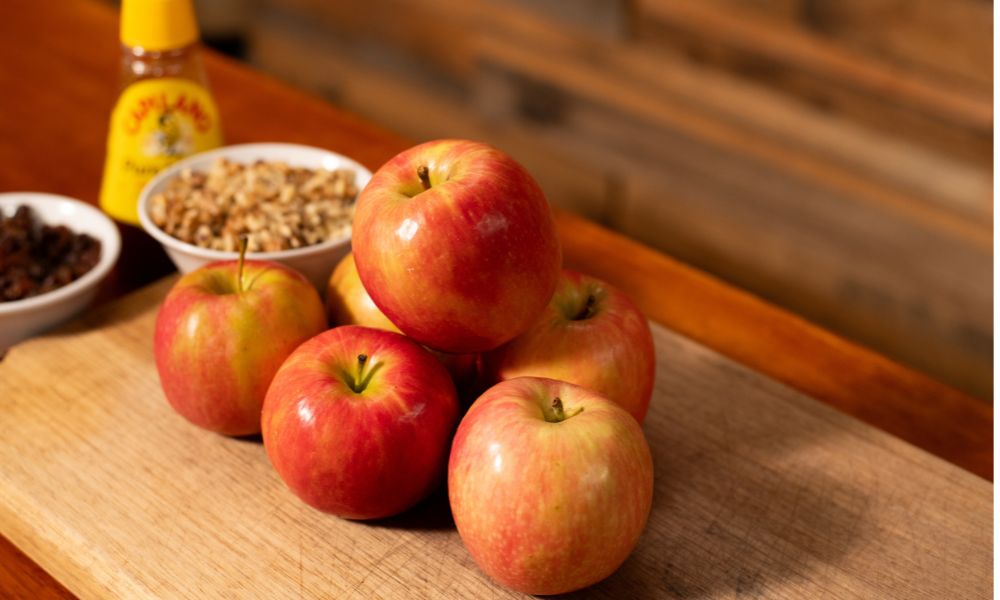 Fresh apples on a wooden board with bowls of chopped nuts and raisins, and a bottle of honey in the background — ingredients for baked apples.