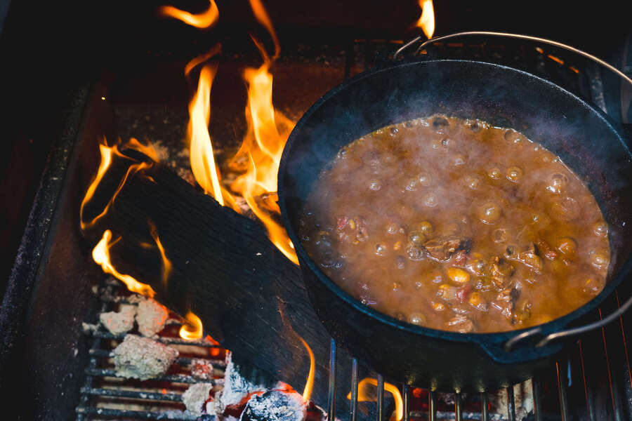 Cast iron pot of beef stew simmering over open flames on a BBQ grill with wood and charcoal burning beneath.