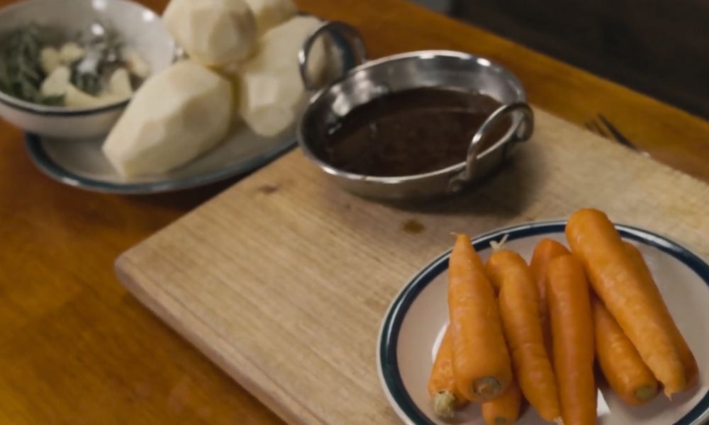 Carrots, peeled potatoes, herbs and a bowl of gravy prepared on a wooden board for cooking.