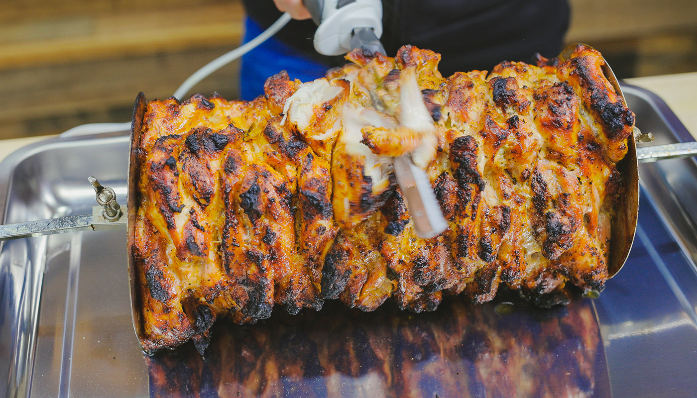 Electric carving knife slicing the outer layer of golden, charred gyro meat on a rotisserie skewer