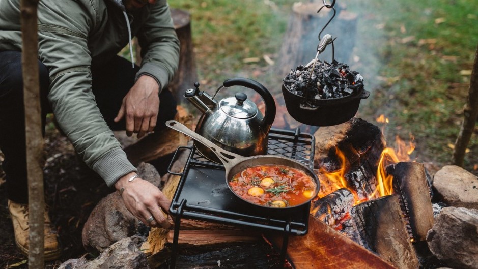 This image shows a man sitting in front of a fire while out camping, cooking on Cast iron cookware combo set