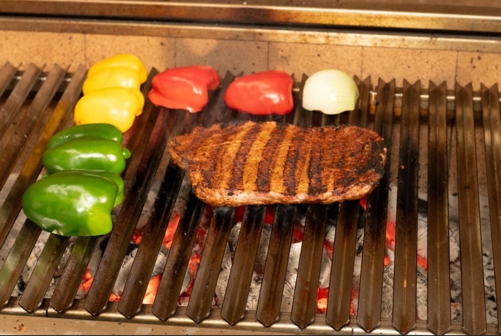 Grilled skirt steak with visible char marks cooking on a Flaming Coals Parrilla BBQ, surrounded by green, yellow, and red capsicum halves and a white onion over glowing charcoal.