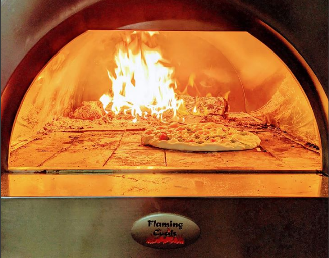 This image shows a fire burning inside of a woodfired pizza oven with a pizza being cooked in it.