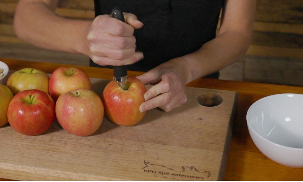 Person using an apple corer to remove the core from a fresh apple on a wooden cutting board, preparing apples for baking.
