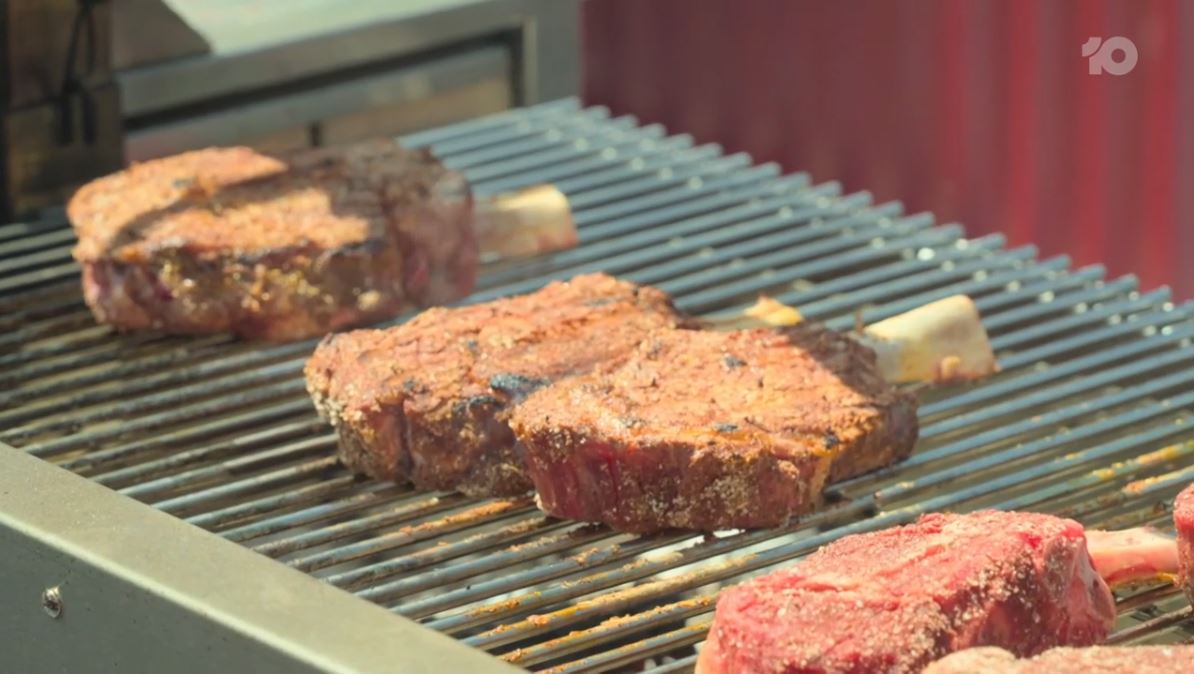 Close-up of double-bone pork chops and rib steaks cooking on a Parrilla grill, with a golden sear on the meat and grill marks visible.