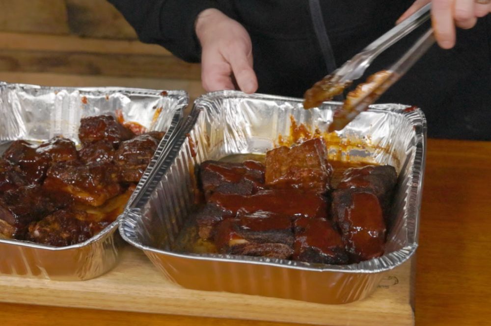 A person uses tongs to glaze smoked pork belly bites in foil trays. The pork is coated in a rich, sticky barbecue sauce, ready for the final caramelisation stage on the BBQ.          Ask ChatGPT