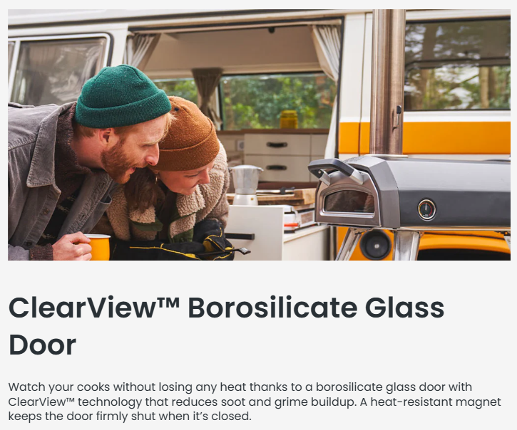 A couple bundled in warm clothing leans in to watch food cooking inside the Ooni Karu 2 Multi-Fuel Pizza Oven, which features a ClearView™ borosilicate glass door. The oven is set up outdoors beside a retro camper, showing how the glass door allows clear visibility while keeping heat sealed in.