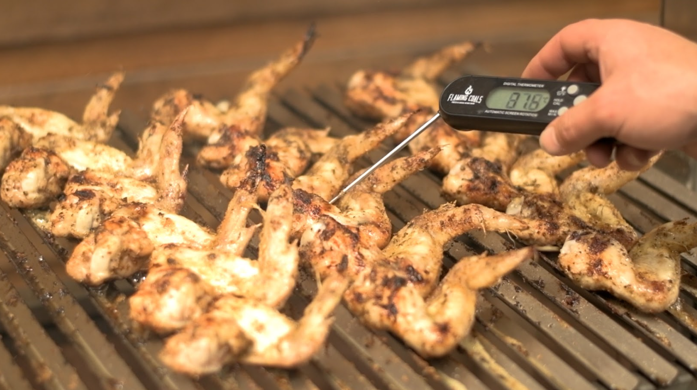Chicken wings cooking across wide stainless steel V channel parrilla grill surface with digital thermometer, showing plenty of cooking space without going freestanding.