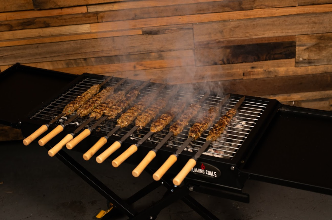 Raw minced meat kebabs cooking on flat skewers over a Flaming Coals Cyprus Spit grill, with smoke rising as they begin to sizzle.