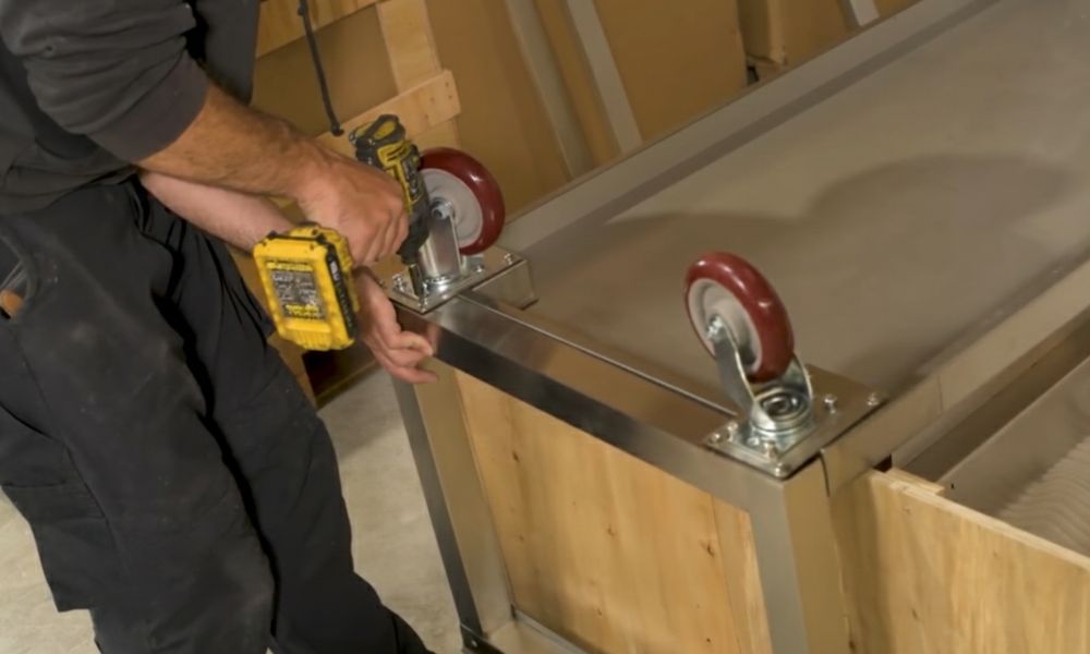 Person attaching heavy-duty caster wheels to the stainless steel Parrilla Argentina grill frame using a power drill.