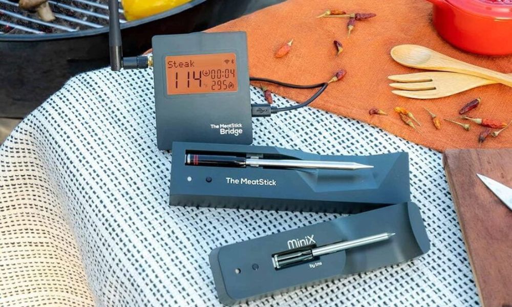 MeatStick WiFi Bridge Set displayed outdoors with digital temperature monitor and two wireless meat probes on a picnic table beside BBQ tools.