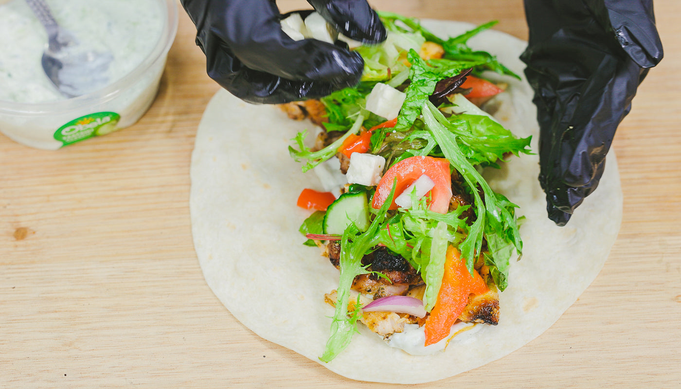 Close-up of pita bread being filled with sliced gyros meat, fresh lettuce, tomato, cucumber, onion, and feta cheese. A person wearing black gloves assembles the wrap on a wooden surface with a bowl of tzatziki sauce nearby.