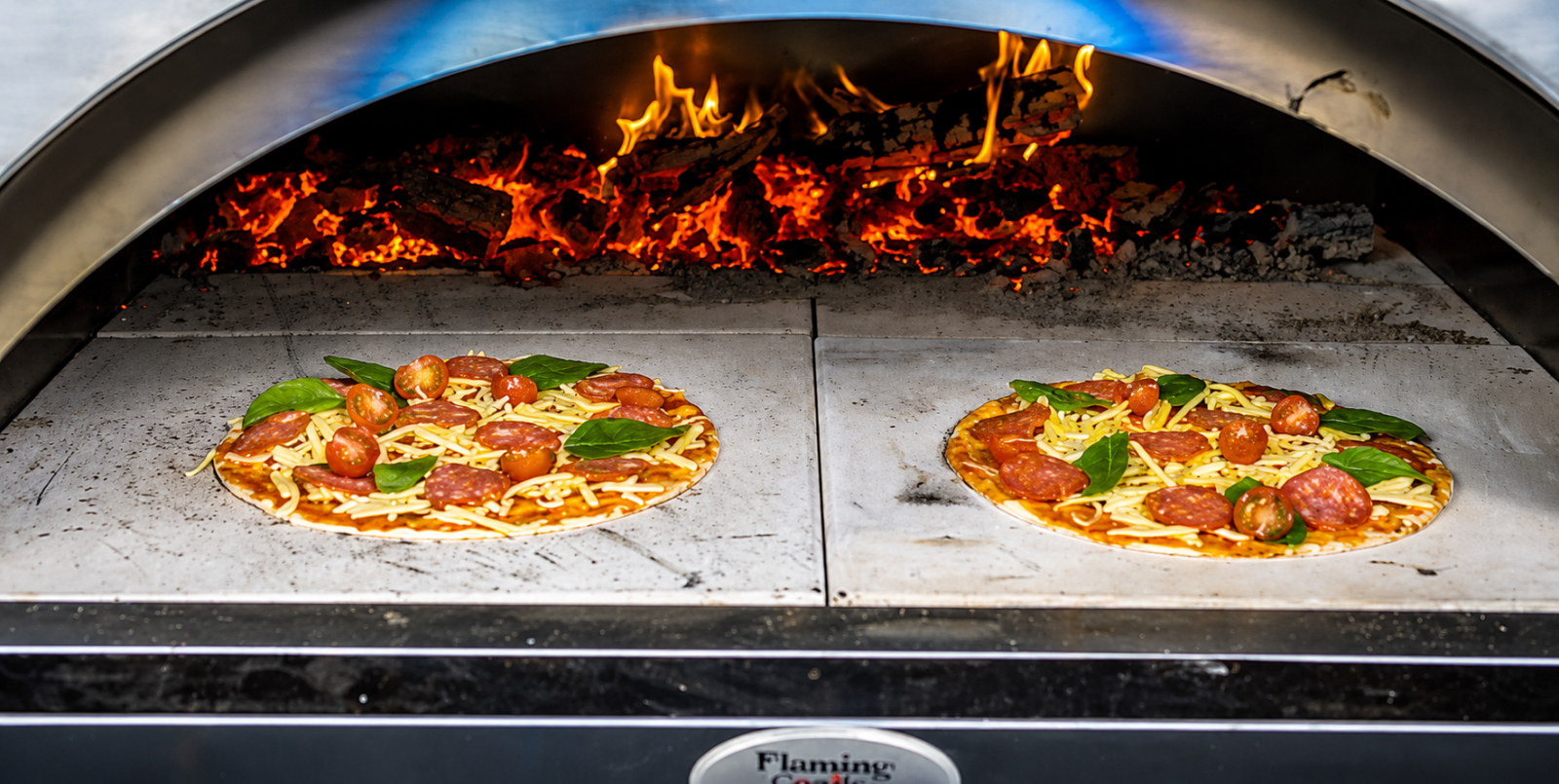 Two pizzas topped with pepperoni, cherry tomatoes, cheese and basil cooking inside a wood fired pizza oven with glowing flames and embers.
