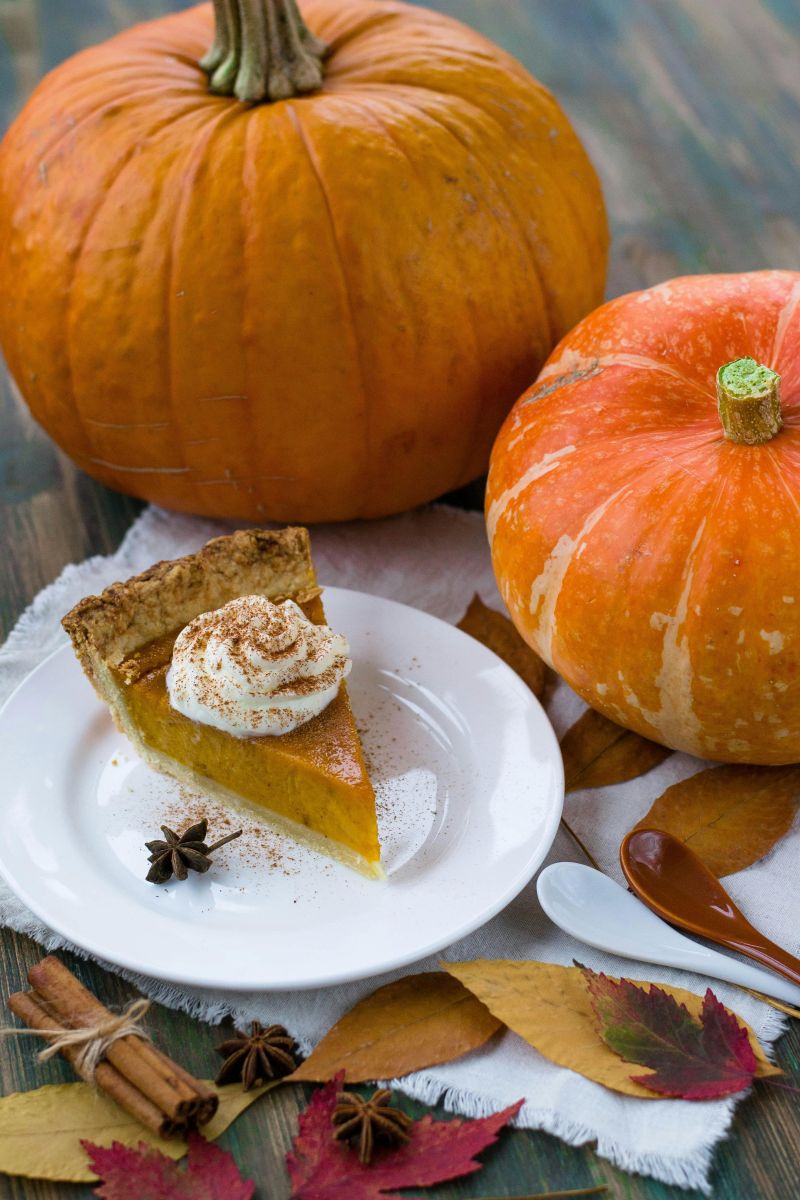 A slice of pumpkin pie topped with whipped cream and sprinkled with cinnamon, served on a white plate next to whole pumpkins and autumn leaves.
