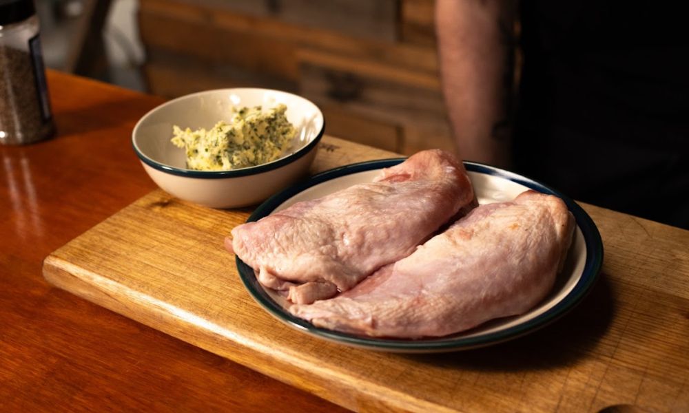 Raw turkey breasts on a plate with a bowl of herb butter on a wooden board.